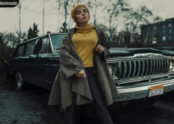 زنی با استایل پاییزی پلیور زرد رنگ پوشیده با اورکت ایستاده و تکیه به یک ماشین شاسی بلند داده.